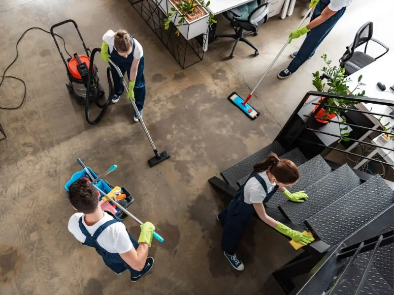 Team of professional cleaners wearing gloves working together to vacuum, mop, and wipe surfaces in a modern office space.