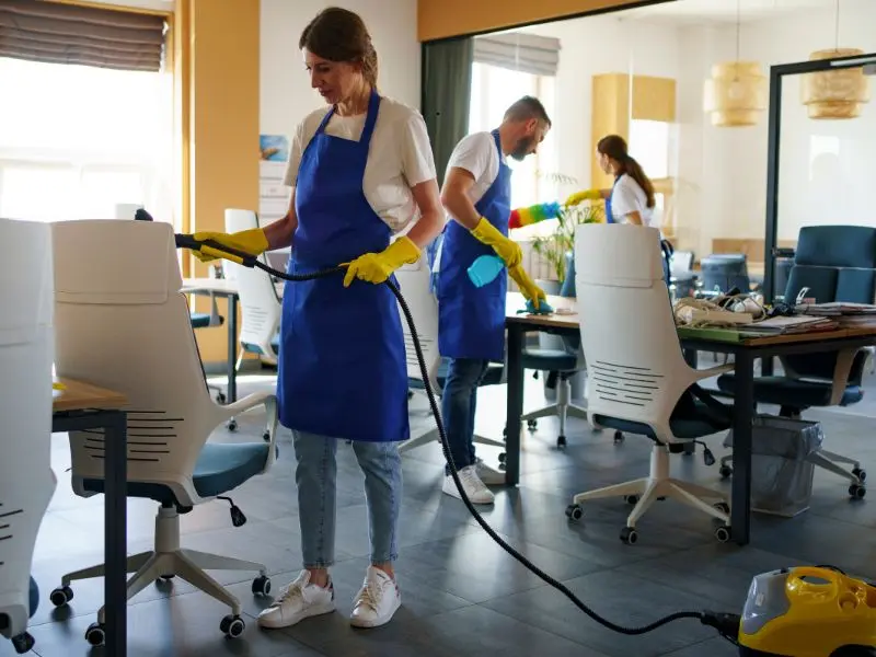 Office cleaning staff in blue aprons and yellow gloves sanitizing chairs, desks, and surfaces in a bright modern workspace.