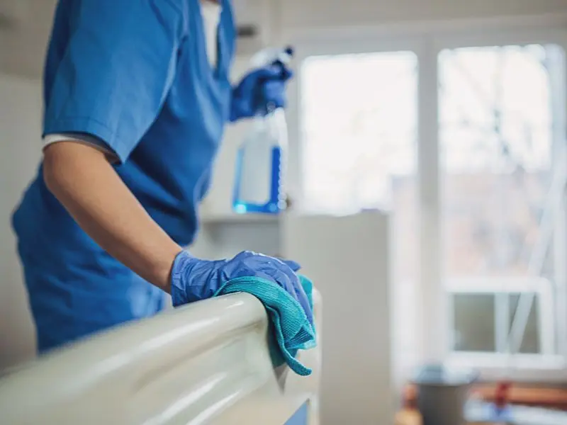 Healthcare worker in blue scrubs disinfecting a hospital bed rail with spray cleaner and cloth to maintain a sterile medical environment.