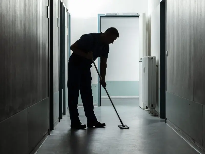 Silhouette of a janitor mopping a dimly lit hallway, ensuring cleanliness and hygiene in a professional building space.