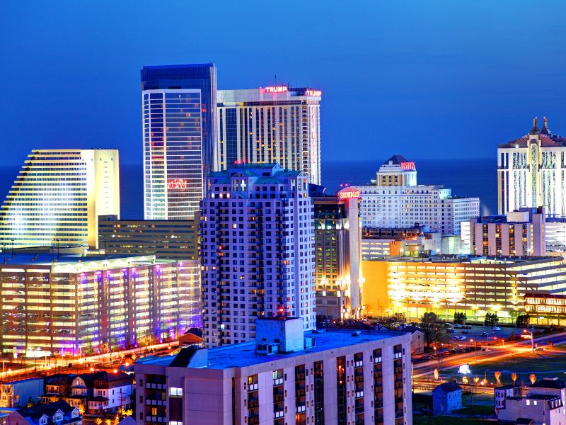 Vibrant night view of New Jersey City’s skyline, featuring brightly lit skyscrapers, casinos, and coastal charm against a deep blue sky.