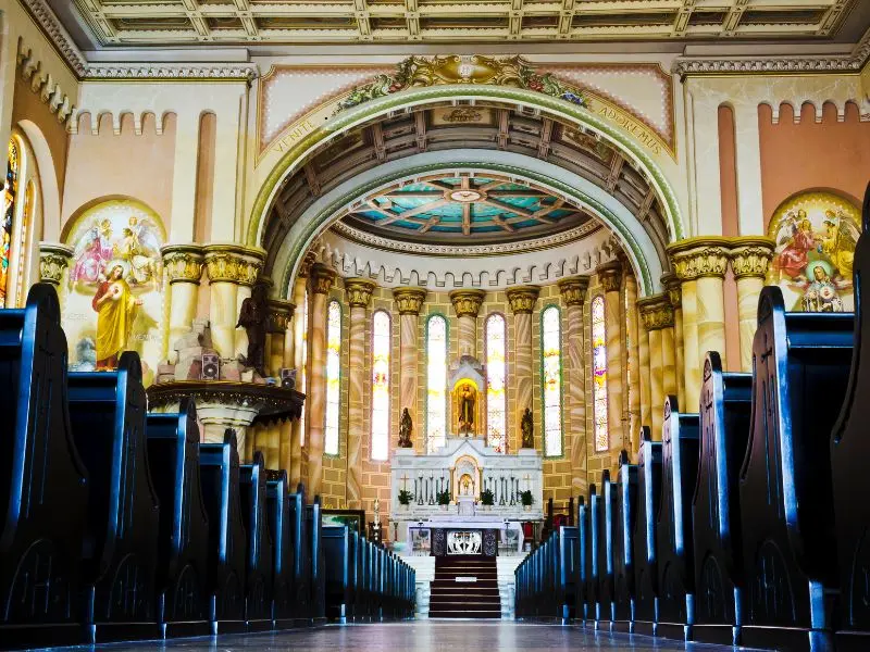 Interior of a historic church with dark wooden pews, colorful stained glass, arched ceiling, and decorative main altar.