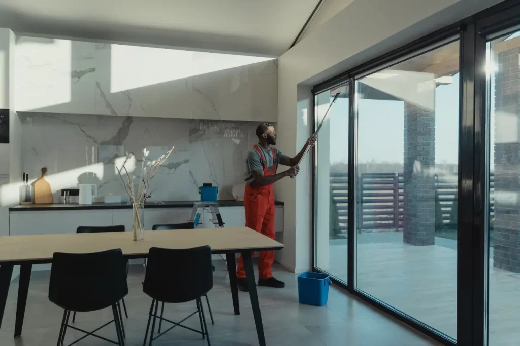 Professional cleaner in uniform using a long-handled tool to wash large glass sliding doors in a modern office workspace with natural light.