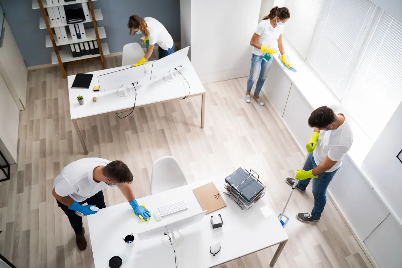 Team of cleaners in uniform delivering office cleaning services by disinfecting desks, windows, and floors for a spotless and hygienic modern workspace.