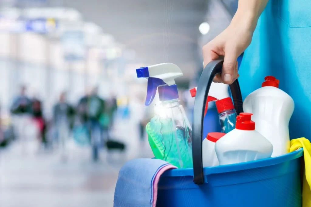Close-up of a person carrying a blue bucket filled with spray bottles, detergents, gloves, and cloths in a busy public area.