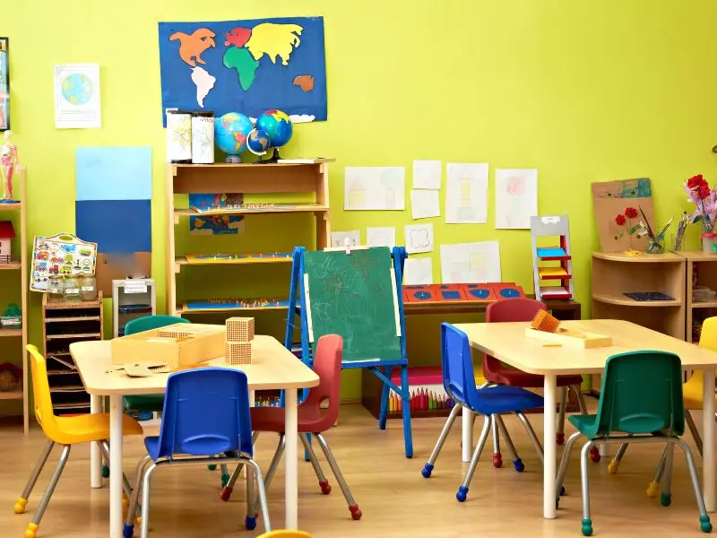 Bright daycare classroom with colorful chairs, clean tables, art supplies, and educational displays on green walls.