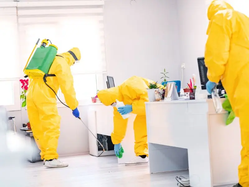 Workers in yellow protective suits disinfecting desks and floors in a corporate office.