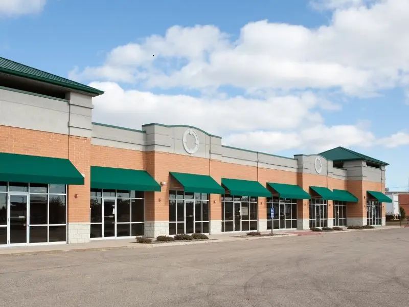 Empty shopping mall with large glass storefront windows and green awnings under a clear sky.