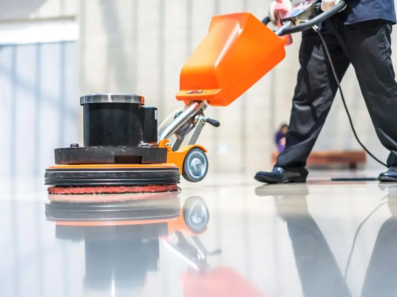 Worker using an orange floor buffer machine to polish commercial flooring.