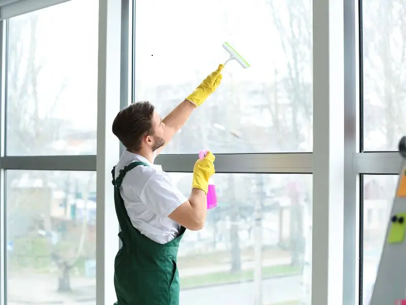 Man in uniform cleaning interior office windows with spray bottle and squeegee in bright workspace.