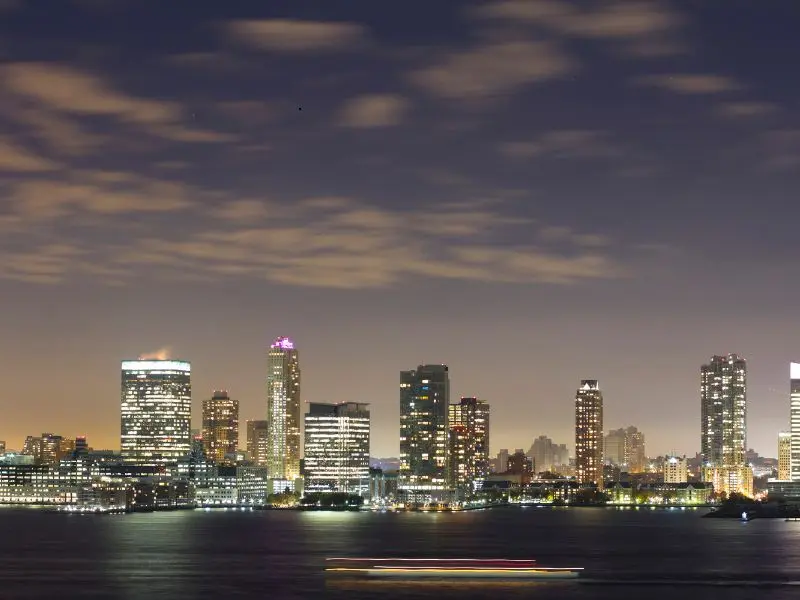 Night view of Jersey City skyline with illuminated skyscrapers and soft reflections over the river.