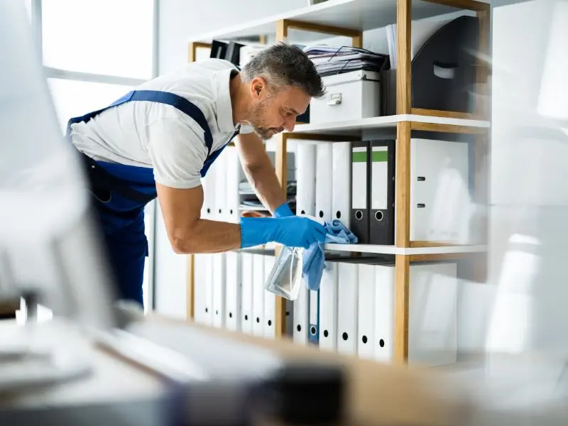 Professional office cleaning technician dusting and organizing file shelves while wearing gloves in a bright workspace with modern storage systems.