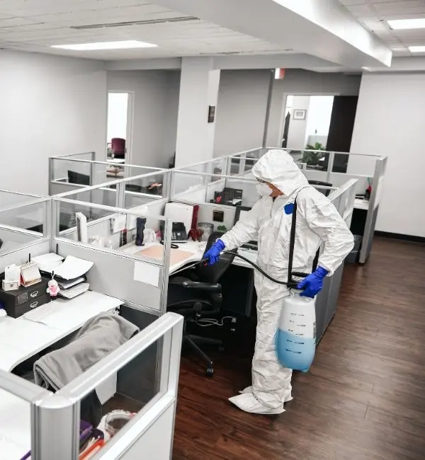 Worker in protective suit disinfects office cubicles with spray equipment, ensuring sanitized workspaces and a safe environment.