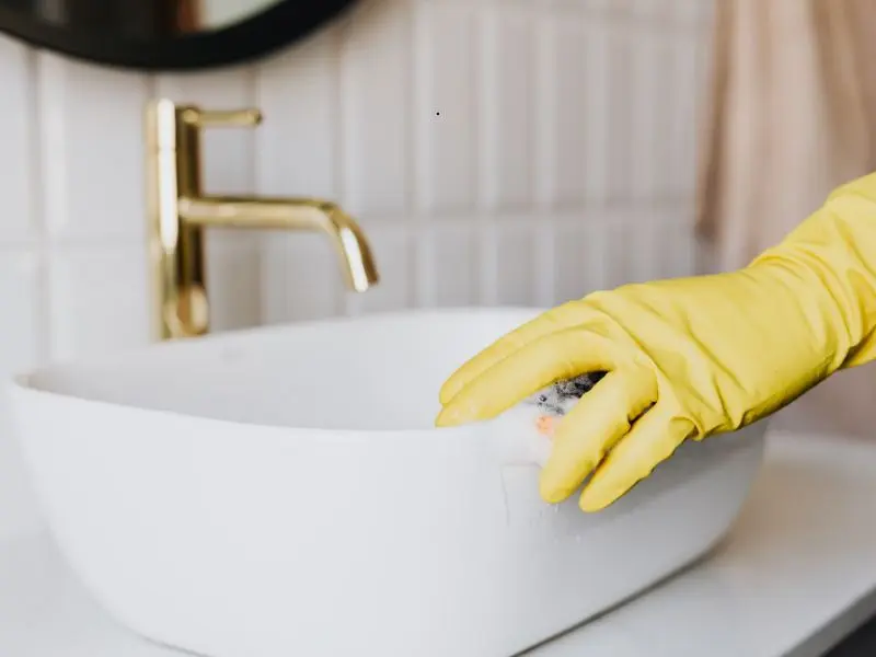 Gloved hand scrubs a modern bathroom sink with soap, highlighting detailed restroom cleaning and sanitization for hygiene and freshness.