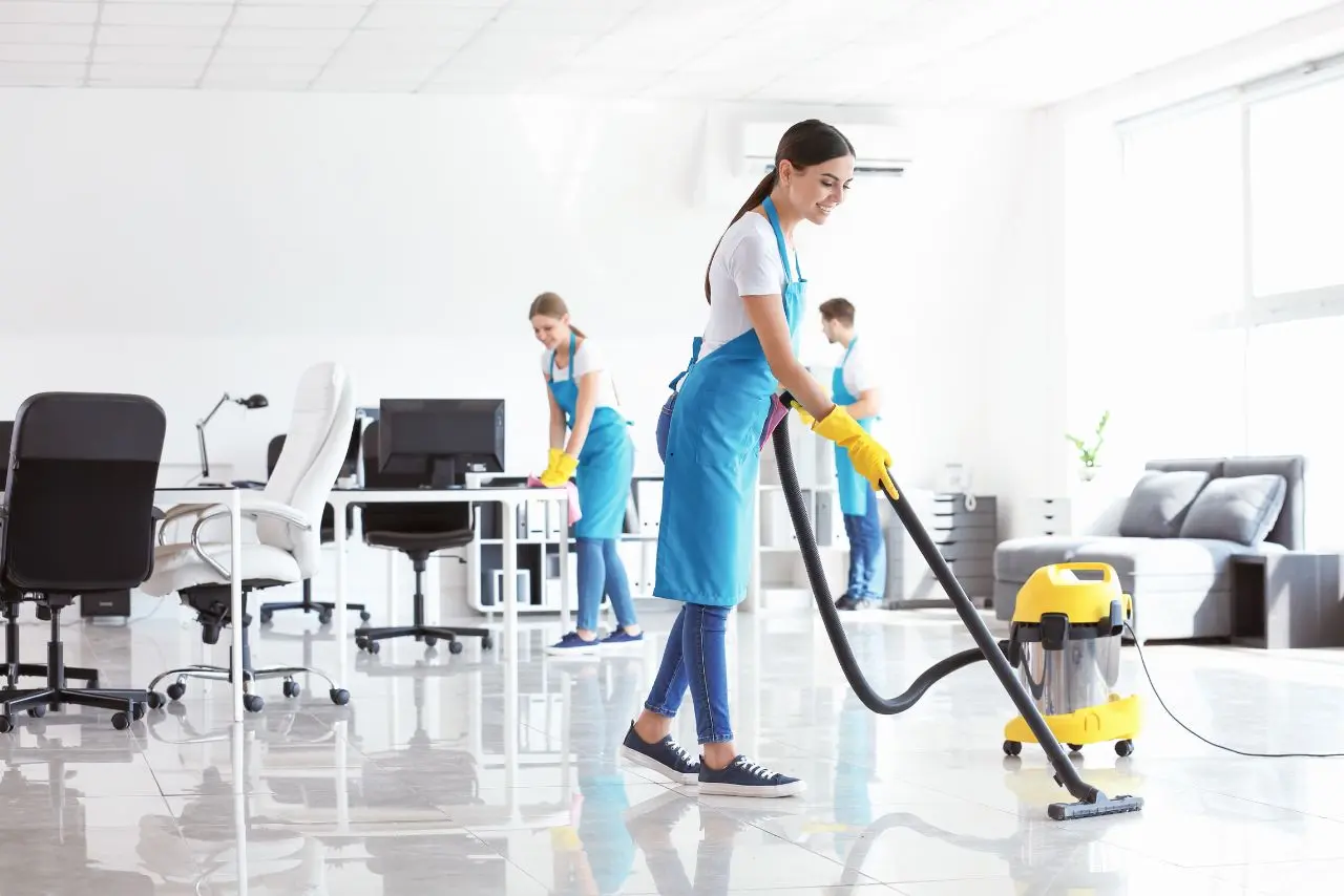 Group of cleaners in uniform using vacuum and supplies to clean a modern, bright, and organized office.