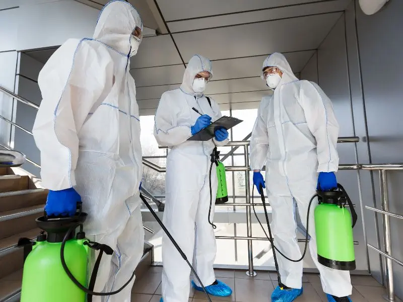 Team in full protective suits and masks holding sprayers, performing sanitation services in a commercial building for health and safety compliance.