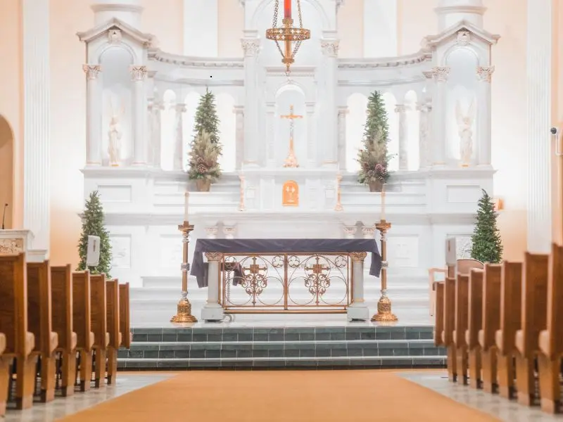 Symmetrical church interior with wooden pews, white marble main altar, gold accents, and soft lighting.