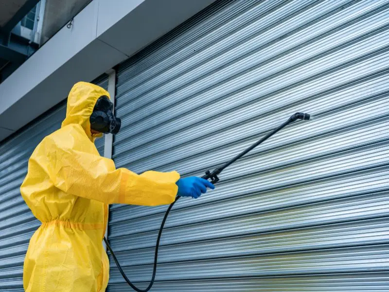 Technician in yellow hazmat suit and mask sprays disinfectant on closed metal shutters, ensuring sanitation and safety in public spaces.