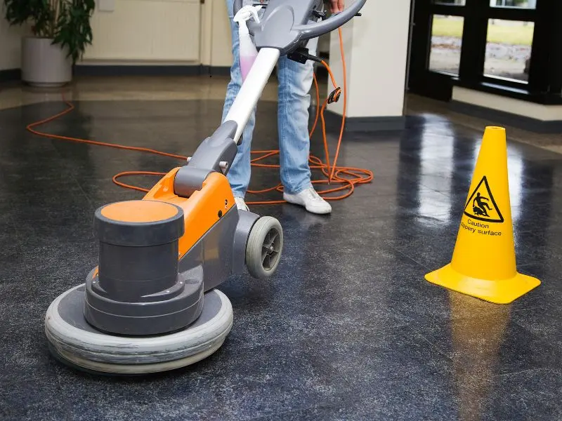 Worker using orange floor buffer on dark surface next to caution sign.