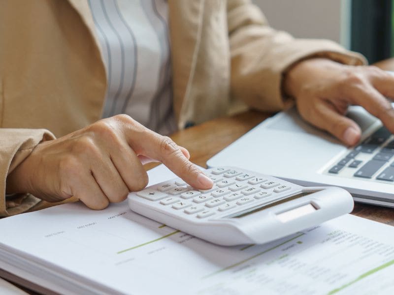 Person using a calculator and laptop to calculate cleaning costs over printed financial documents.