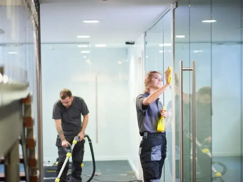 Janitorial services examples with two cleaners, one vacuuming the floor and the other wiping glass office doors