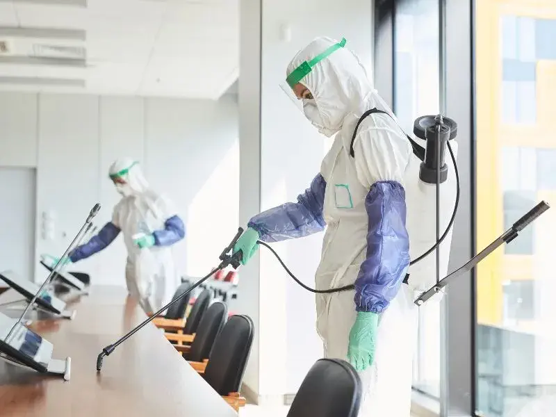 Two professionals in hazmat suits disinfecting desks and screens in a modern office.
