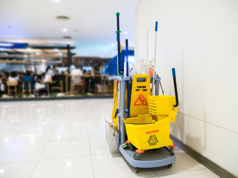 Janitorial day porter cleaning cart with mop, bucket, and supplies in a commercial hallway.