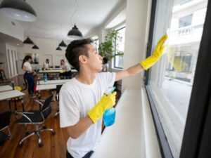 Worker in yellow gloves cleaning a large office window with spray bottle, showing a professional commercial cleaning process indoors