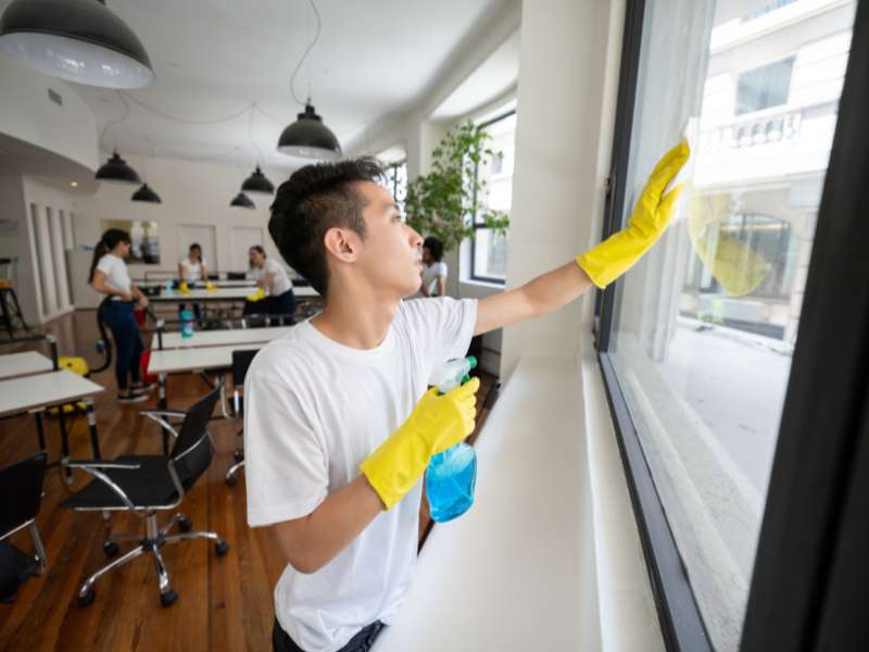 Worker in yellow gloves cleaning a large office window with spray bottle, showing a professional commercial cleaning process indoors
