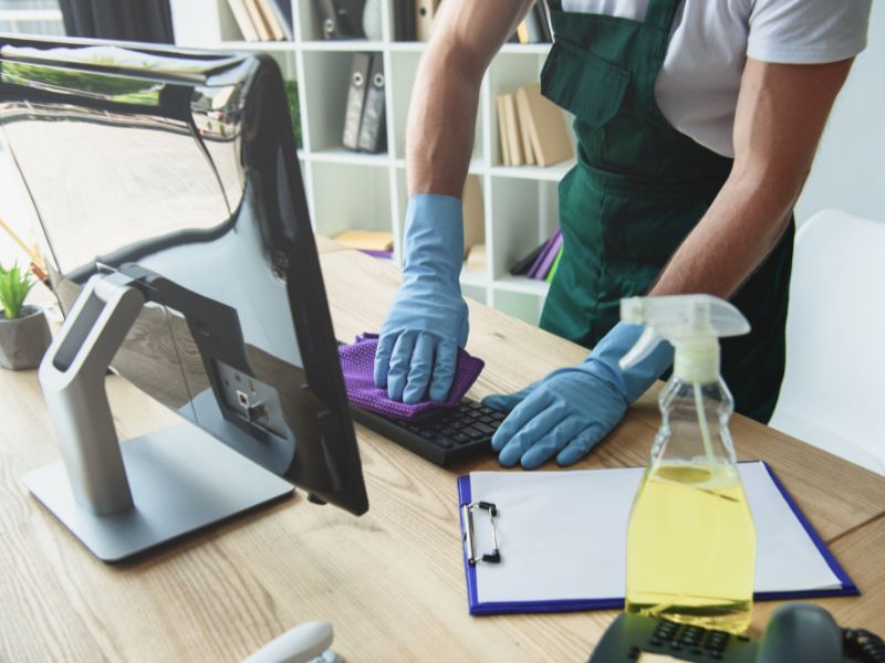 Cleaning professional disinfecting an office desk, keyboard, and workstation with gloves and cleaning solution during routine office maintenance.